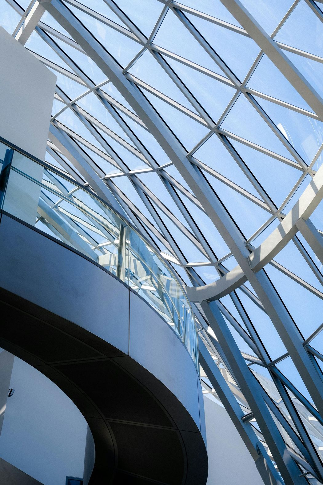 Curved glass atrium ceiling with steel beams and blue sky visible above a modern interior
