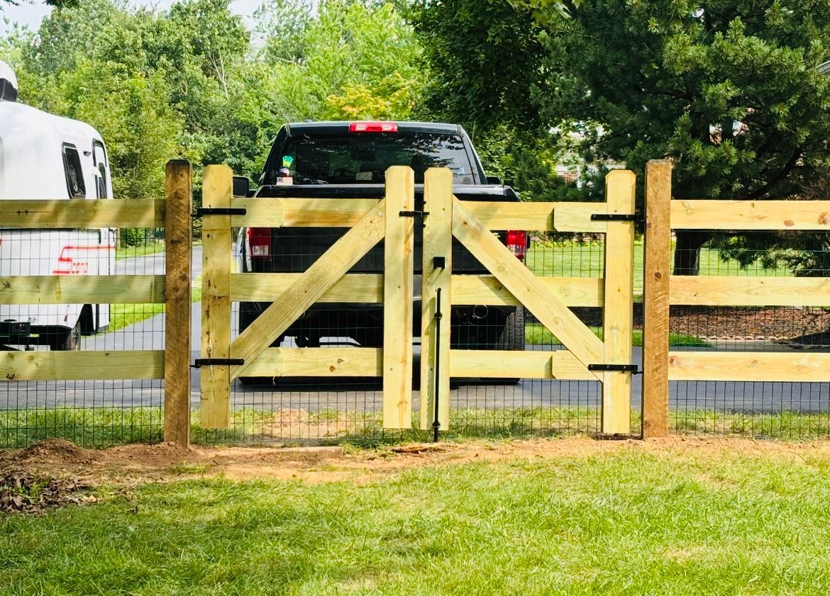 Black pickup truck behind a wooden farm gate in a grassy driveway, with trees in the background