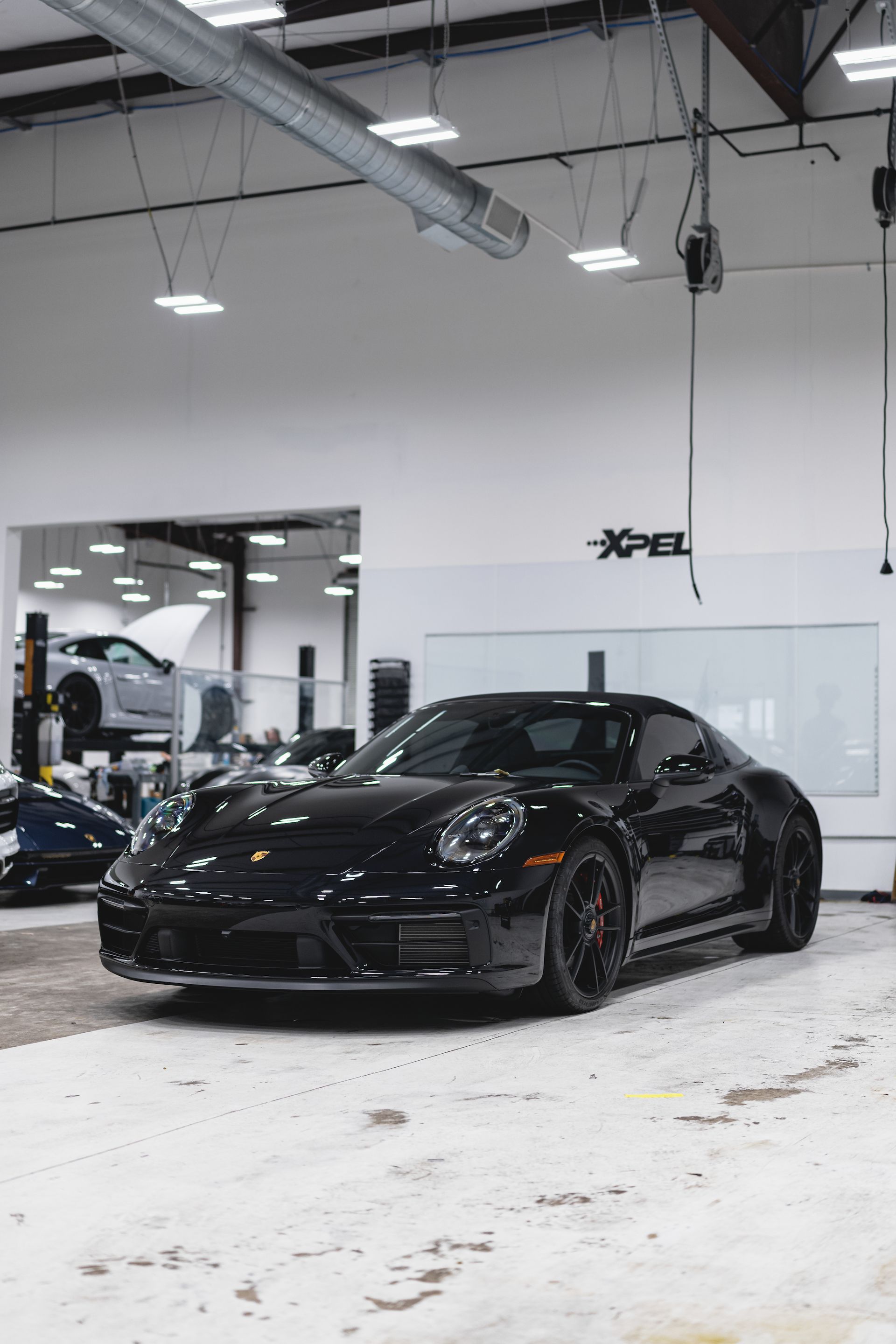 Black Porsche 911 in a bright garage with overhead lights; another car is visible in the background.