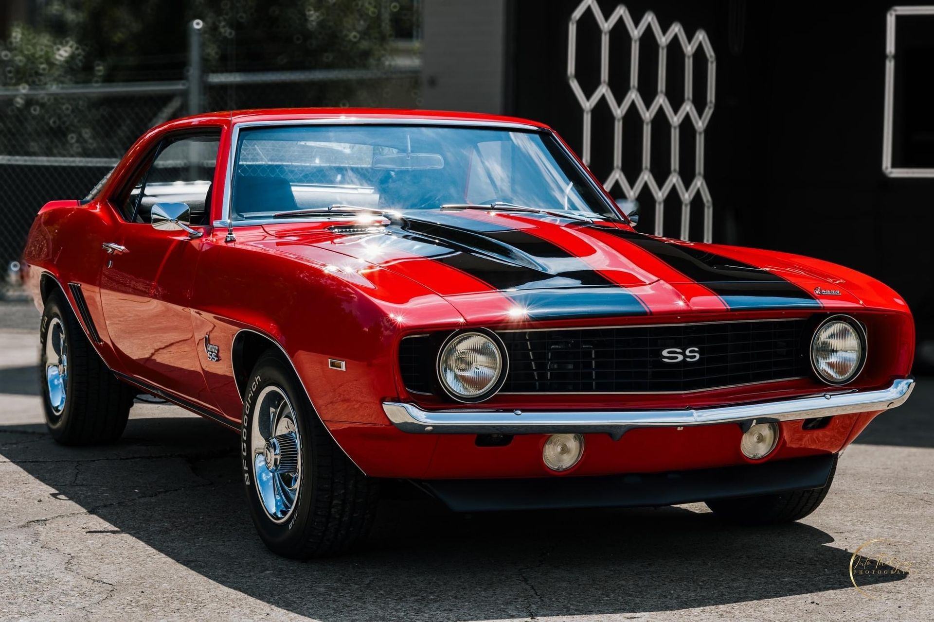 Red and black 1969 Chevy Camaro SS classic car with racing stripes, parked in front of a building.