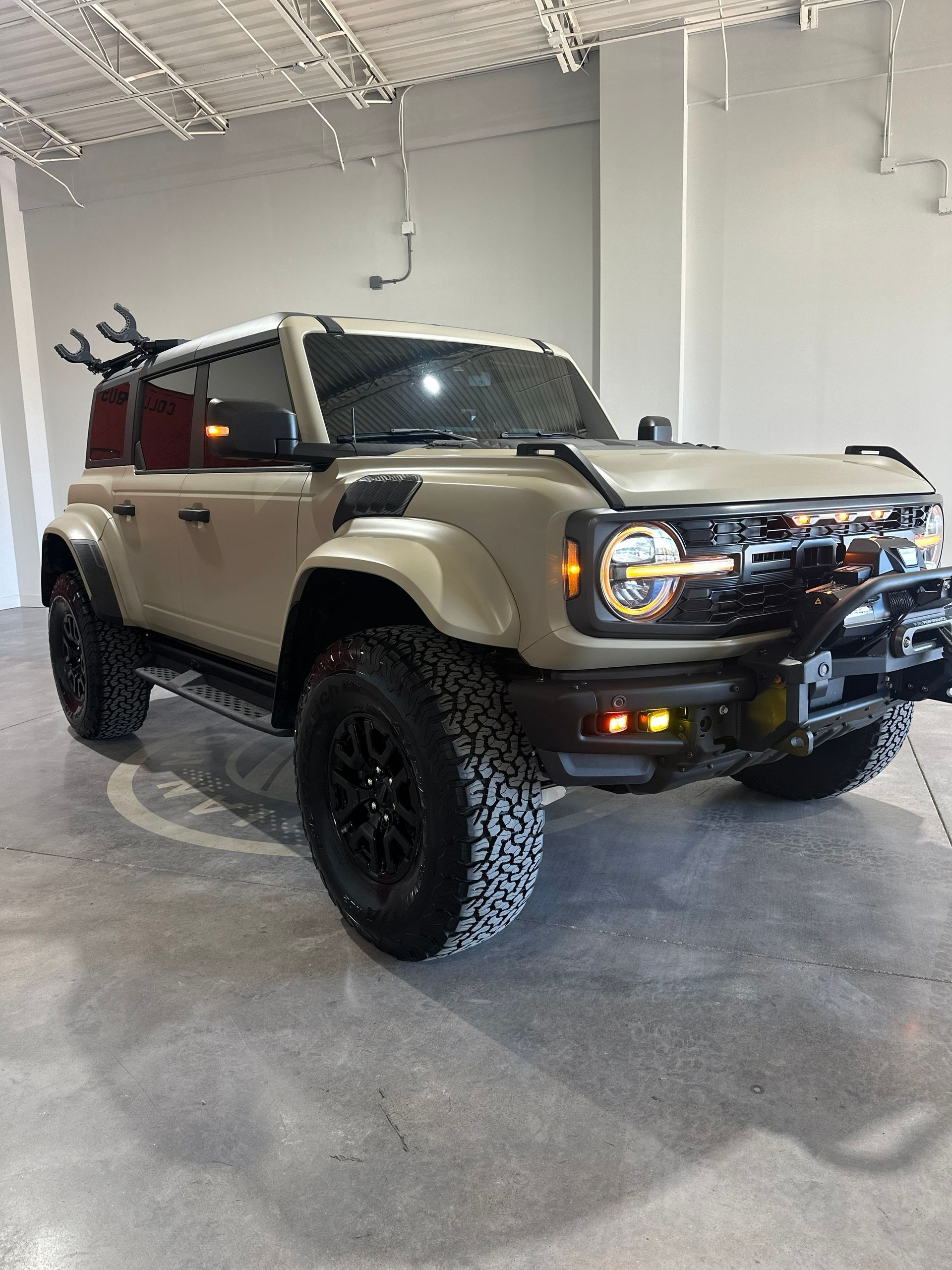 Tan Ford Bronco SUV with black wheels and off-road modifications, parked indoors.
