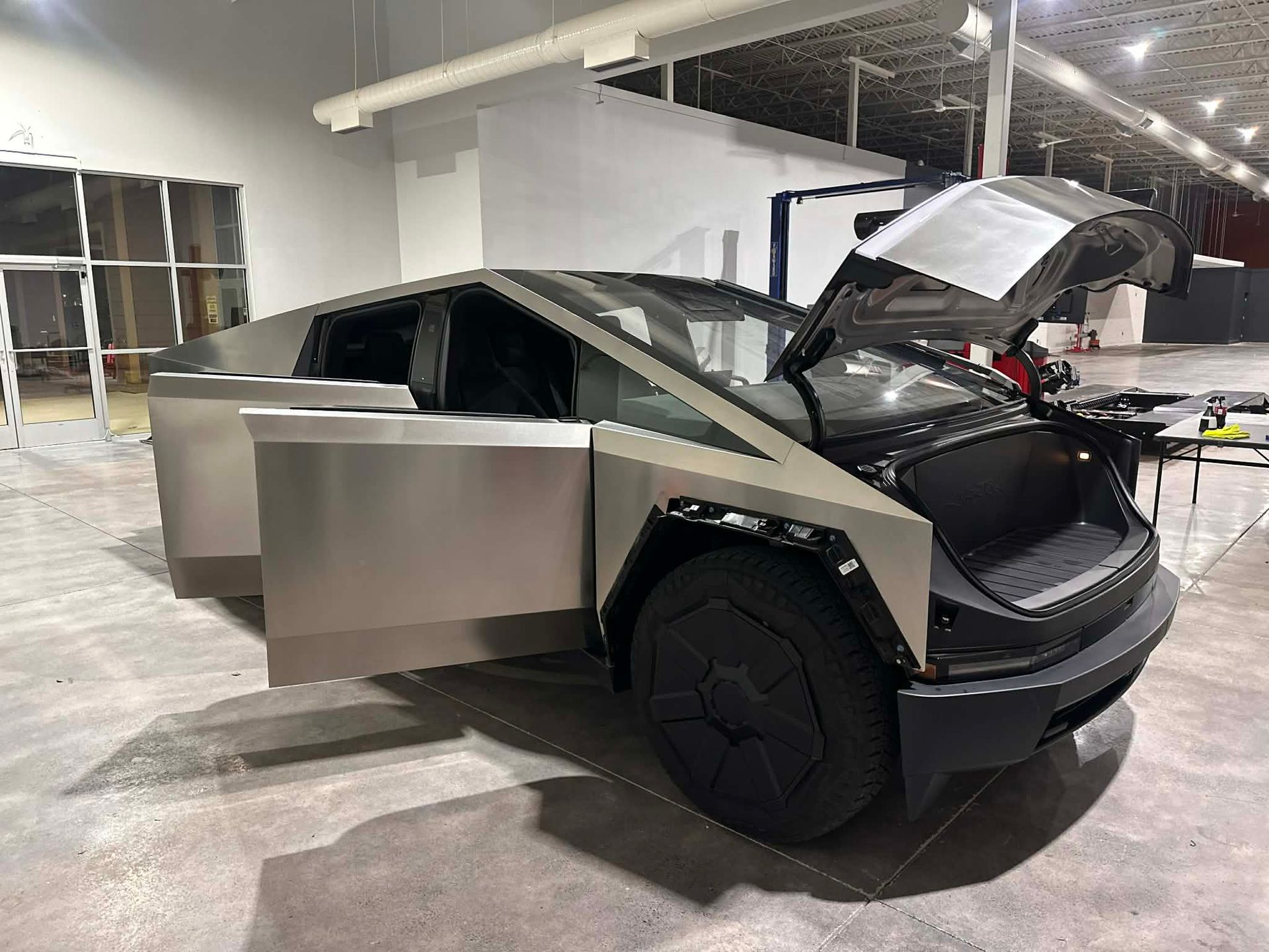 Tesla Cybertruck with doors and front trunk open, indoors. Stainless steel body, black details, gray floor.
