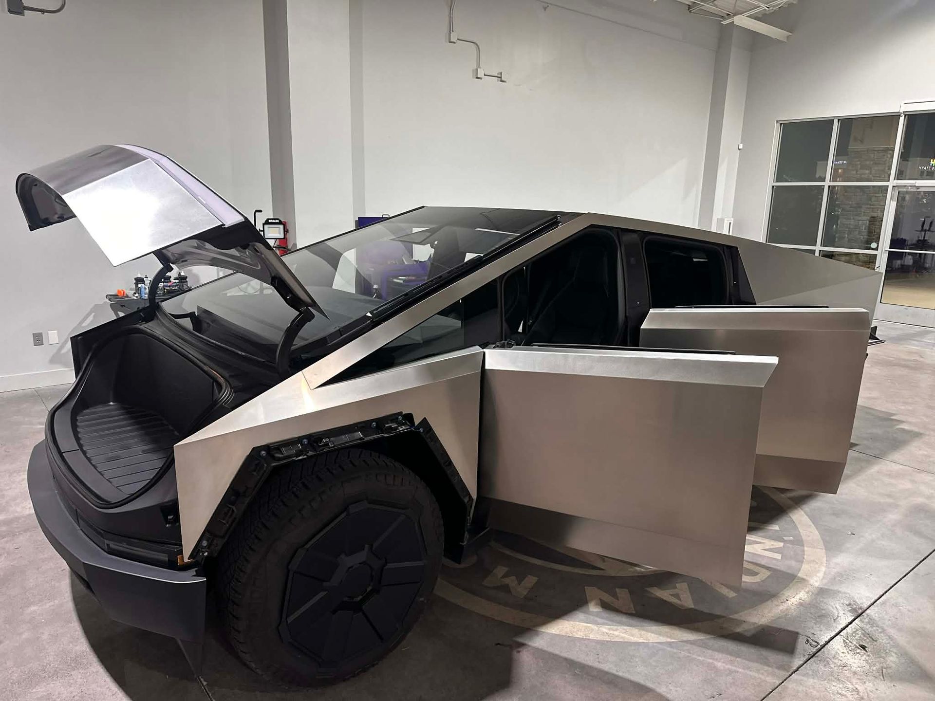 Tesla Cybertruck with doors and front trunk open, indoors. Stainless steel exterior, black wheels.