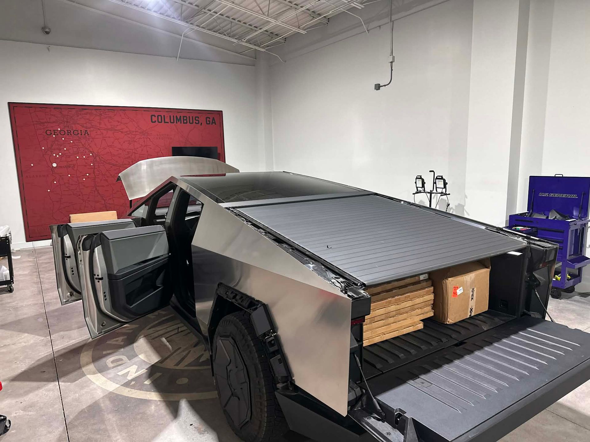 Tesla Cybertruck with open doors and bed, boxes inside, parked in a workshop setting.