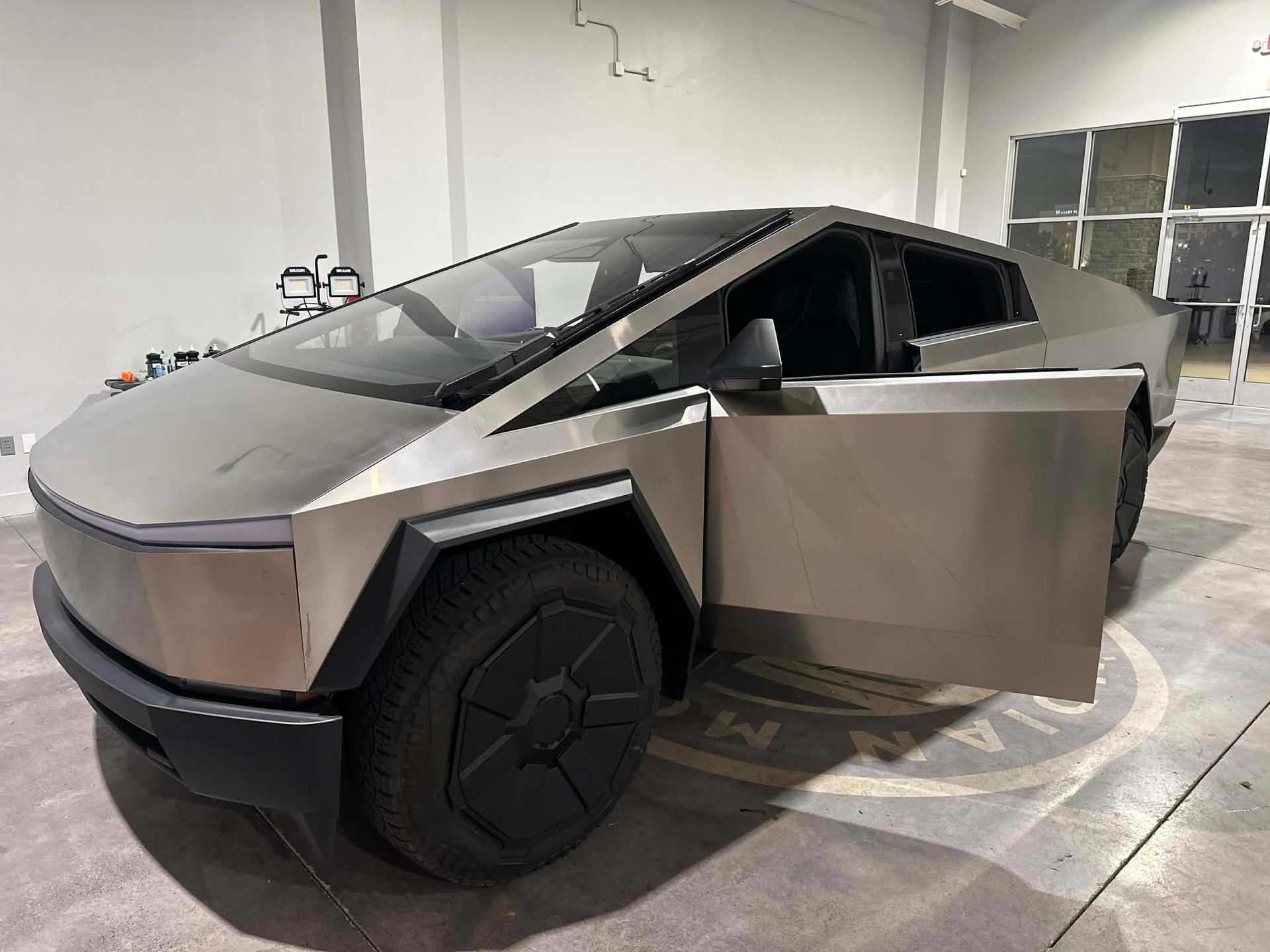 Tesla Cybertruck, silver, in a workshop setting, driver's side door open, black wheels.