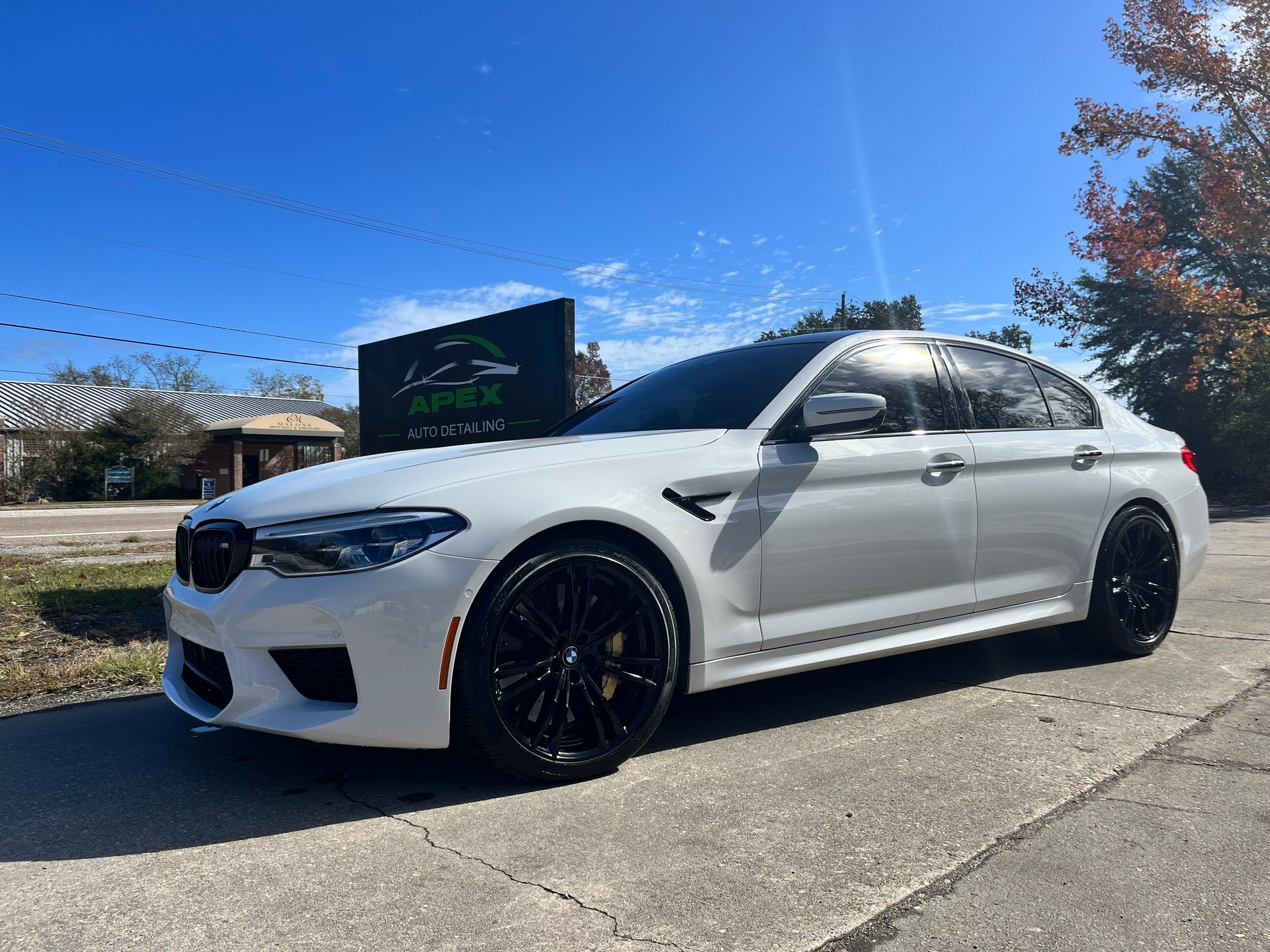 White BMW M5 sedan parked on a paved lot with a business sign in the background. Black wheels. Sunny day.