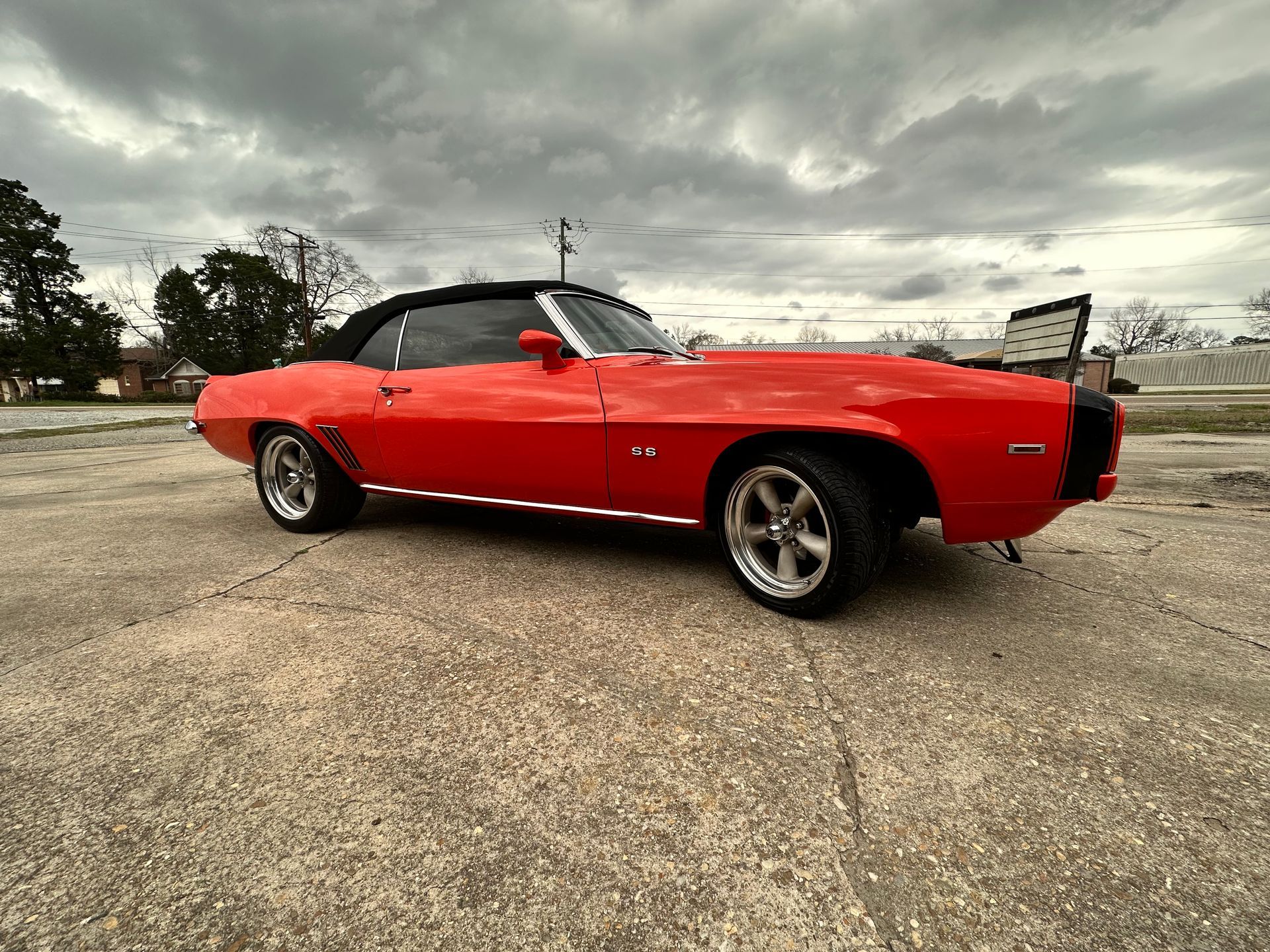 Red convertible Camaro, chrome wheels, black top, parked on gravel. Overcast sky.