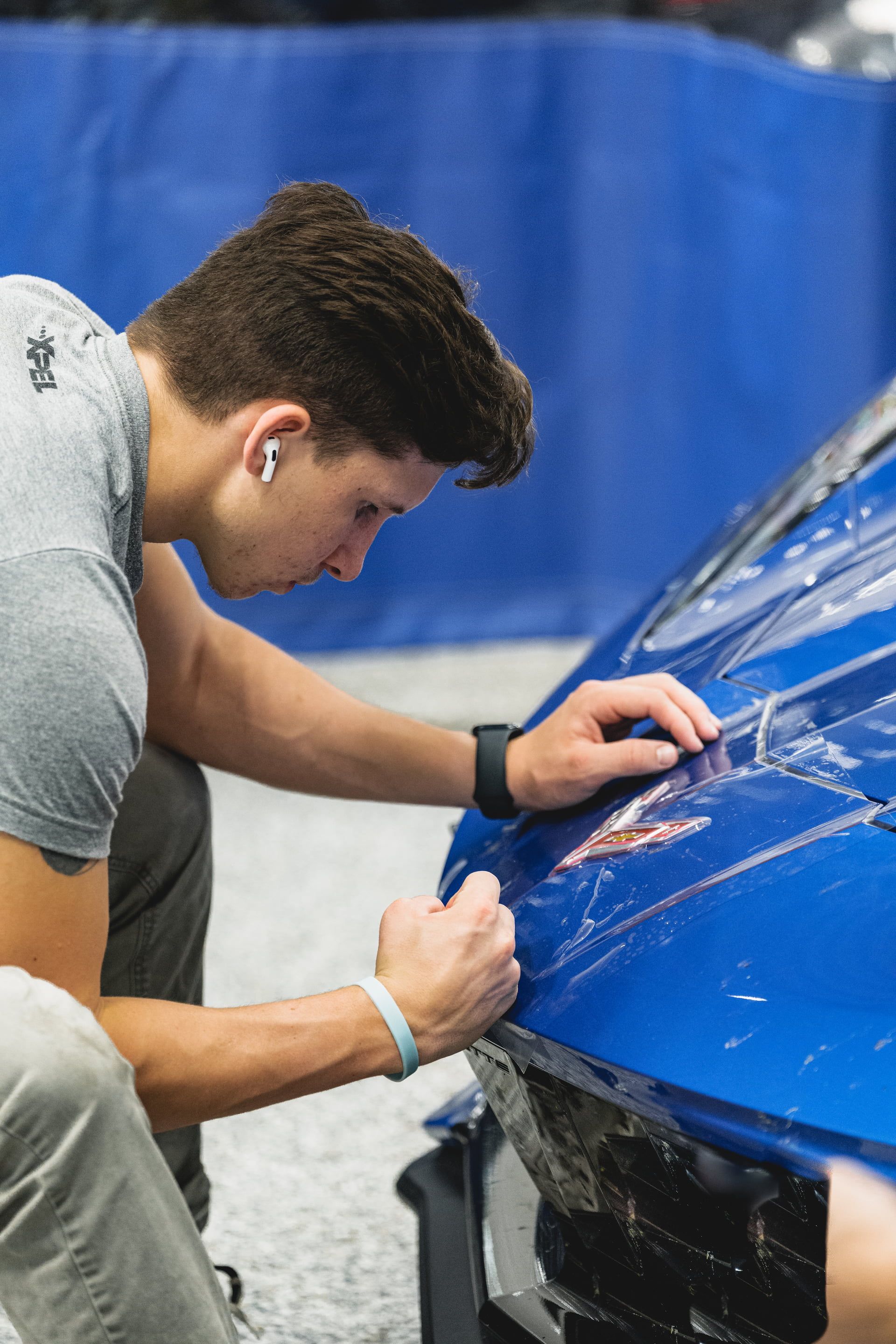 Man applying film to blue car's front. Indoors, focused, hands at work.