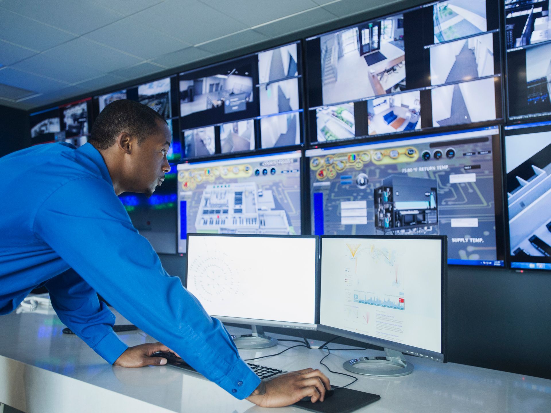 A man is working on a computer in front of a wall of monitors.