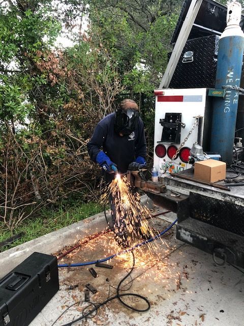 Person welding metal, sparks flying. Outdoors with equipment and trees.