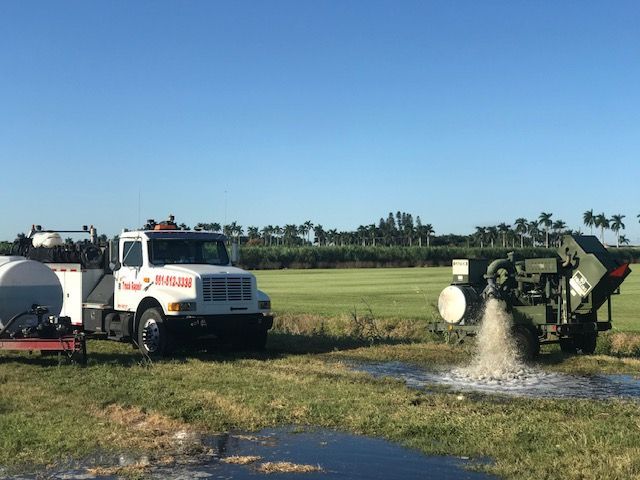 Truck and machinery on field releasing water or foam. Blue sky and palm trees in background.