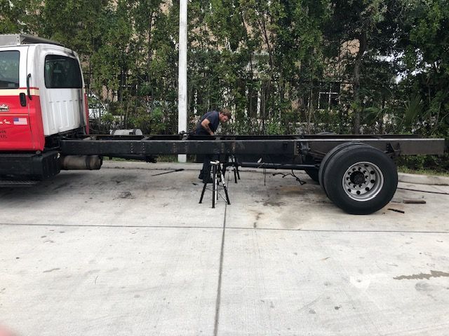 A person works on a truck frame outdoors. The truck is white and red, with a dark frame and two rear tires.