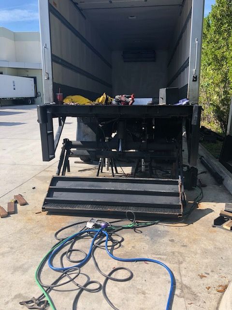 Open truck with a lift gate, tools, and hoses in front. Outdoors, on concrete.