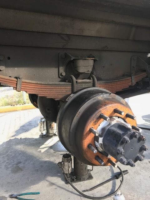 Truck axle with rust-colored leaf spring, black drum brake, and a jack.