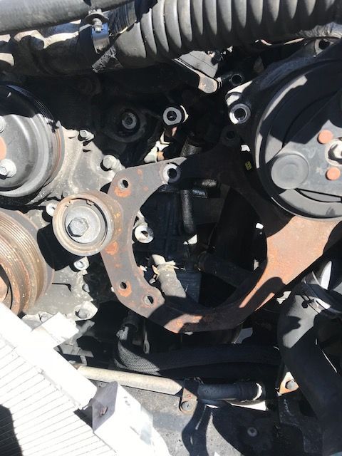 Rusty metal engine bracket with surrounding pulleys and hoses in a car engine bay.