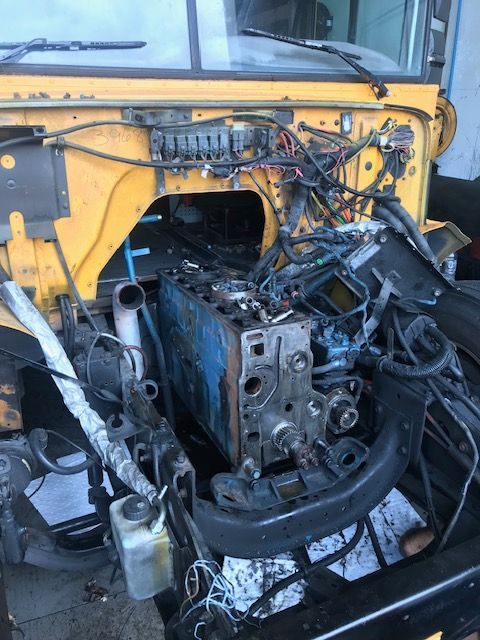 A disassembled school bus engine compartment, showing the engine block, wiring, and metal frame. Yellow and blue hues.