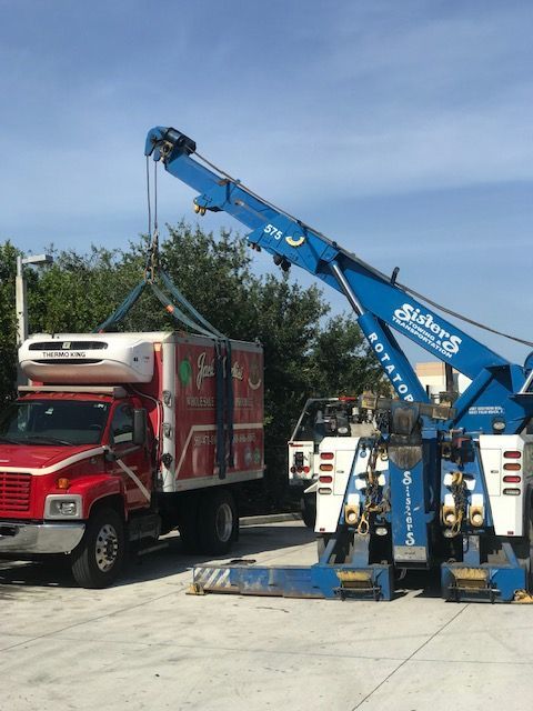 A red box truck being lifted by a blue tow truck with a refrigerated unit on top.