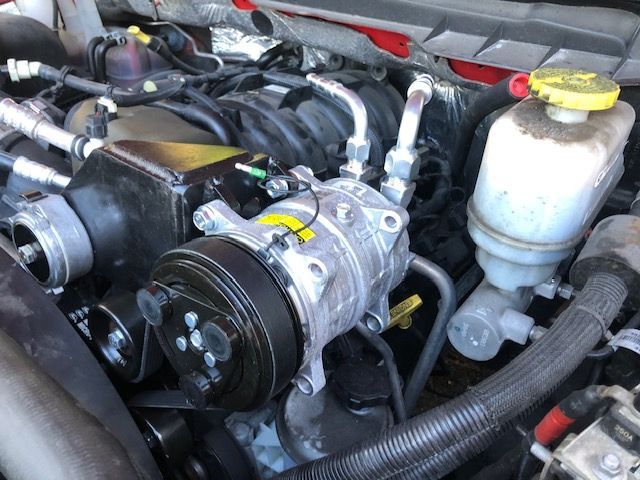 Engine bay of a vehicle showing an air conditioning compressor and other engine components.