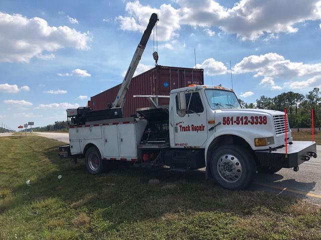 A tow truck uses a crane to lift a shipping container on a roadside under a cloudy sky.