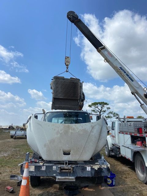 Crane lifting an engine out of a truck's open hood, outdoors under a blue sky.