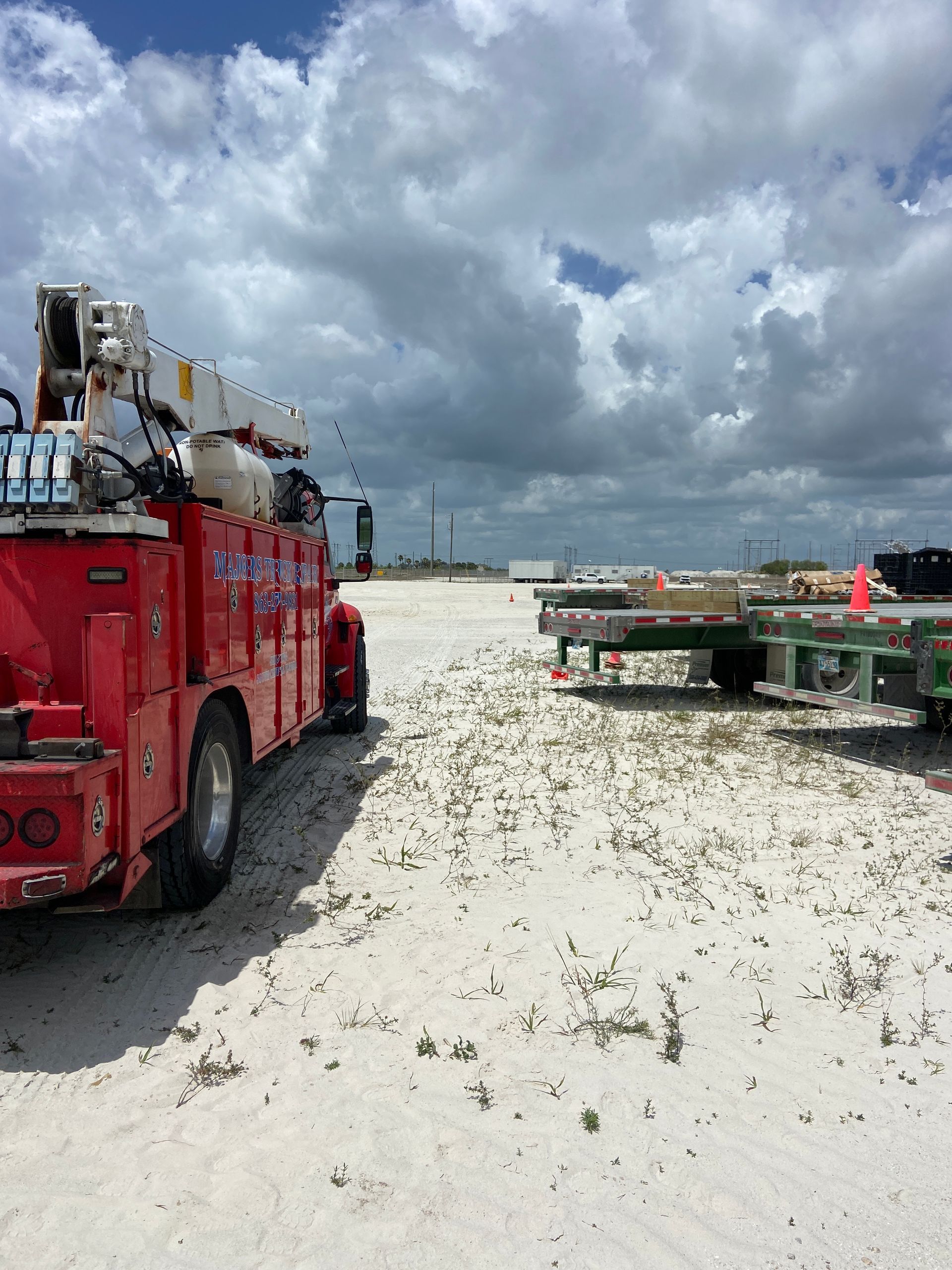 Red truck with crane parked on sandy ground, trailers in background, cloudy sky.