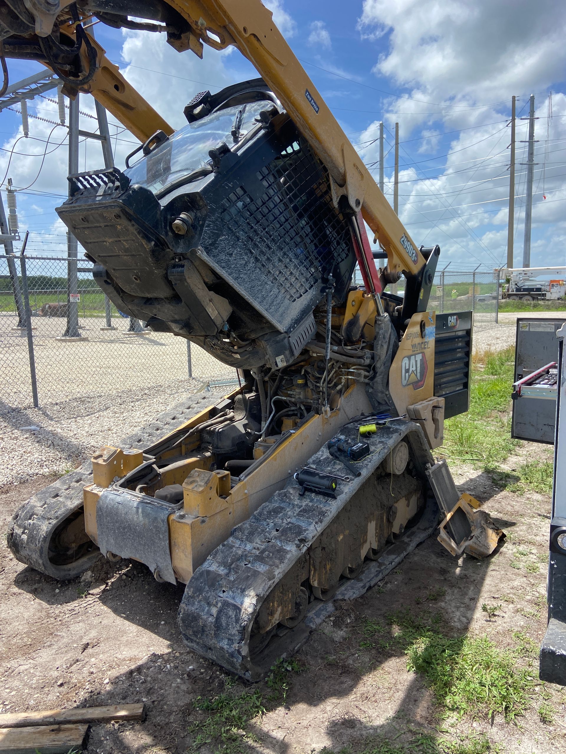 Yellow and black skid steer with the hood open, tracks on the ground, near power lines.