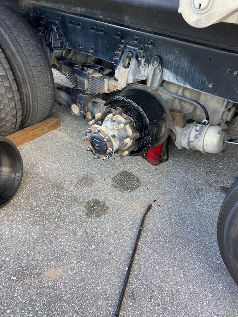 Black semi-truck axle and tire on pavement. A wooden block and wrench are present.