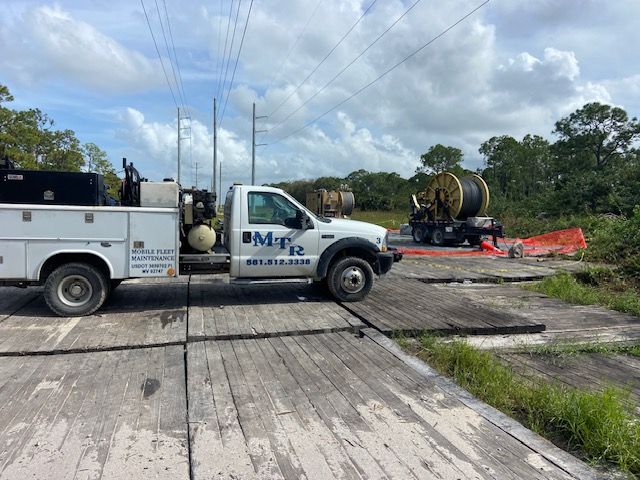 Utility truck parked on wooden planks near power lines and a cable reel. Cloudy sky.
