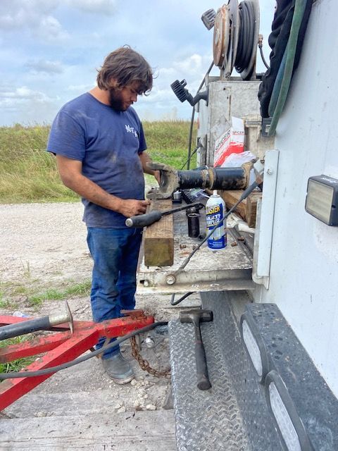 Man working on machinery near a truck. He wears a blue shirt and jeans, standing outside on a cloudy day.