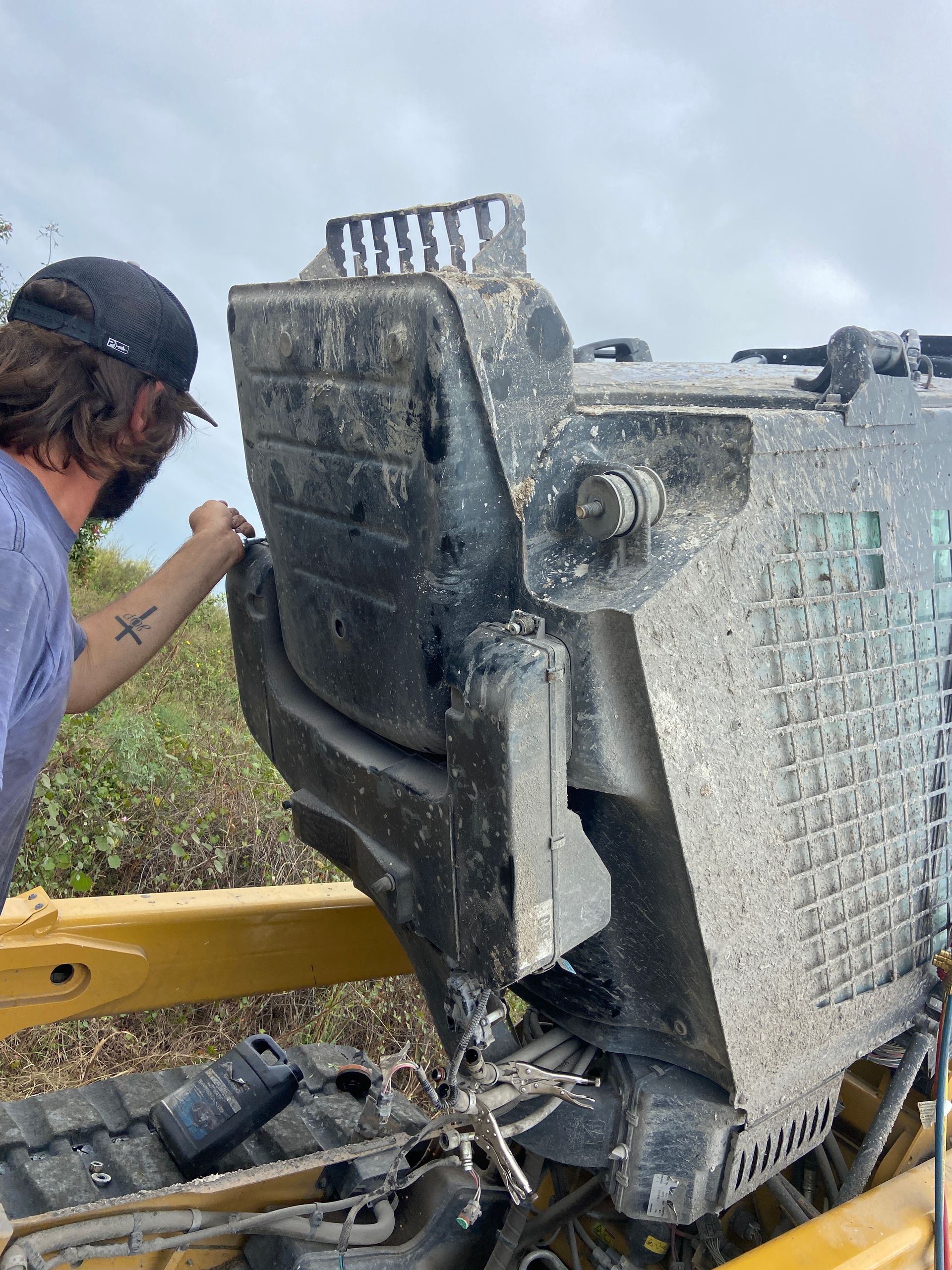 Man examining a dirty, black piece of heavy machinery, on a yellow tracked vehicle, outdoors.