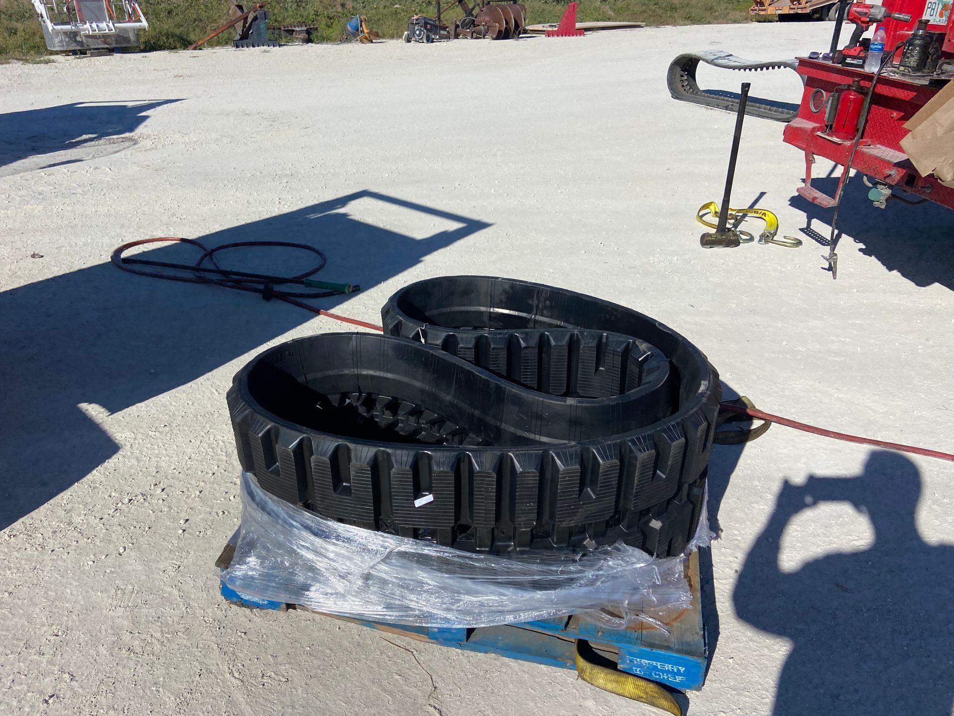 Black rubber track coiled on a pallet, outdoors on concrete with equipment in the background.