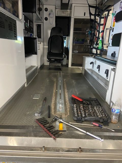 Interior of an ambulance being worked on, tools scattered on floor. Black chair in the center, white cabinets on sides.