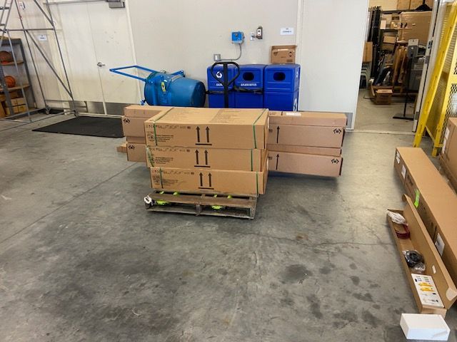 Boxes on a pallet in a warehouse. Blue recycling bins, a cart, and other items are in the background.