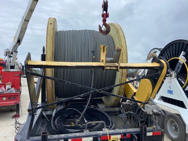 Large spool of gray cable mounted on a trailer, suspended by a crane hook outdoors.