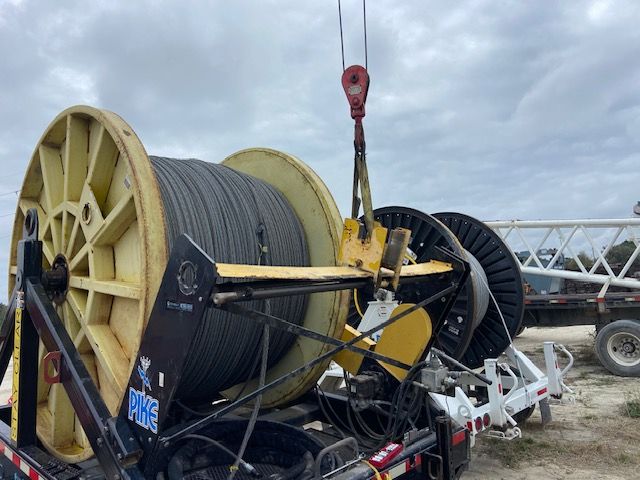 A large spool of black cable being lifted by a crane hook in an outdoor setting.