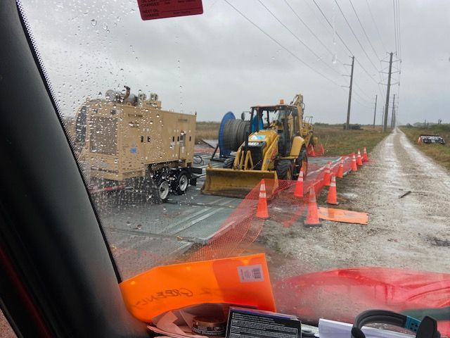 A work scene with a backhoe, trailer, and orange safety cones on a muddy road under power lines.