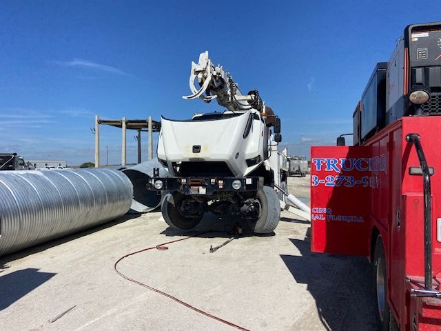 White concrete pump truck at a construction site. Red service truck on the right, large metal pipes on the left.