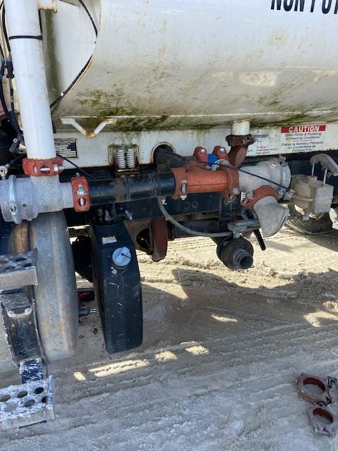 Side view of a white tank truck with pipes, valves, and a pressure gauge visible.