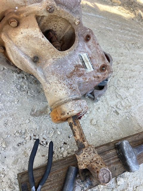 Rusty, industrial machine component with a metal shaft, pliers, and a hammer on a wooden plank, outdoors.