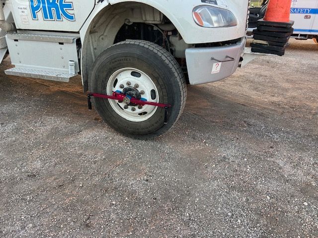 A truck's front wheel is secured with a red strap. The truck is parked on gravel.