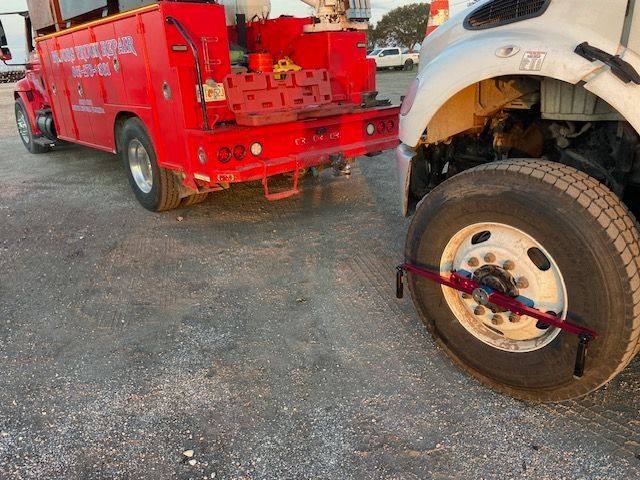 Red tow truck towing a white truck with a damaged front, wheel fixed.