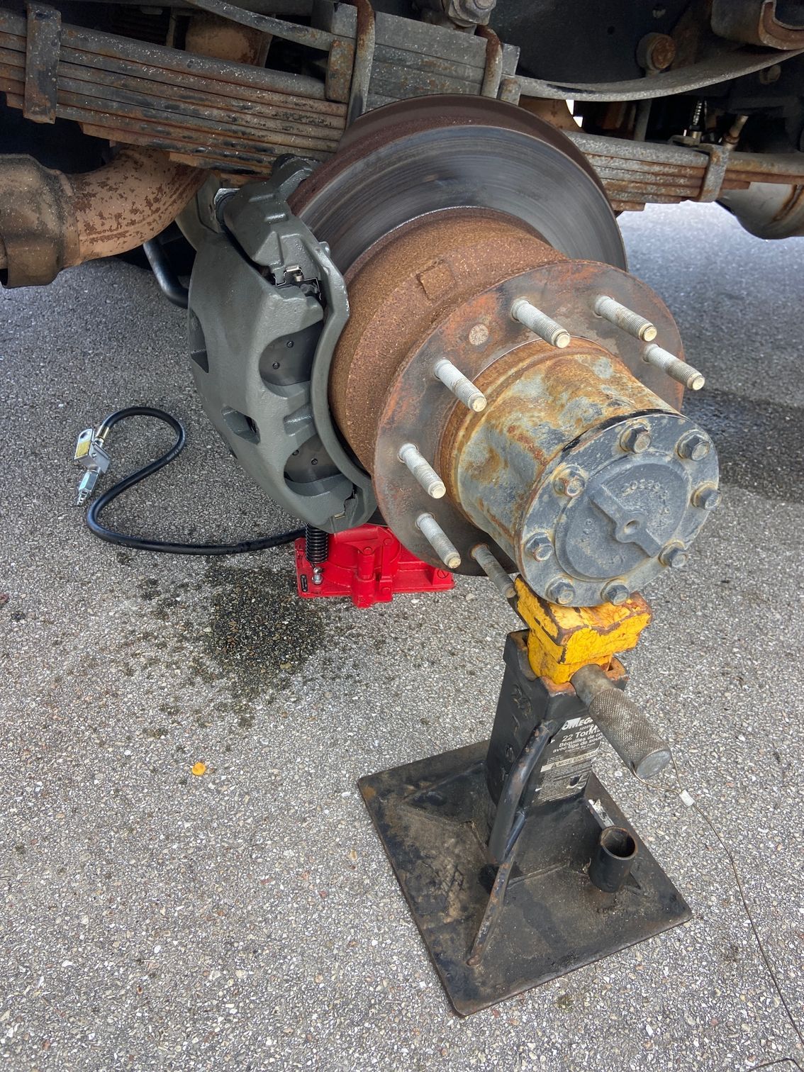 Truck wheel assembly on jack stand; rusty brake rotor, gray caliper, red jack, metal leaf springs.