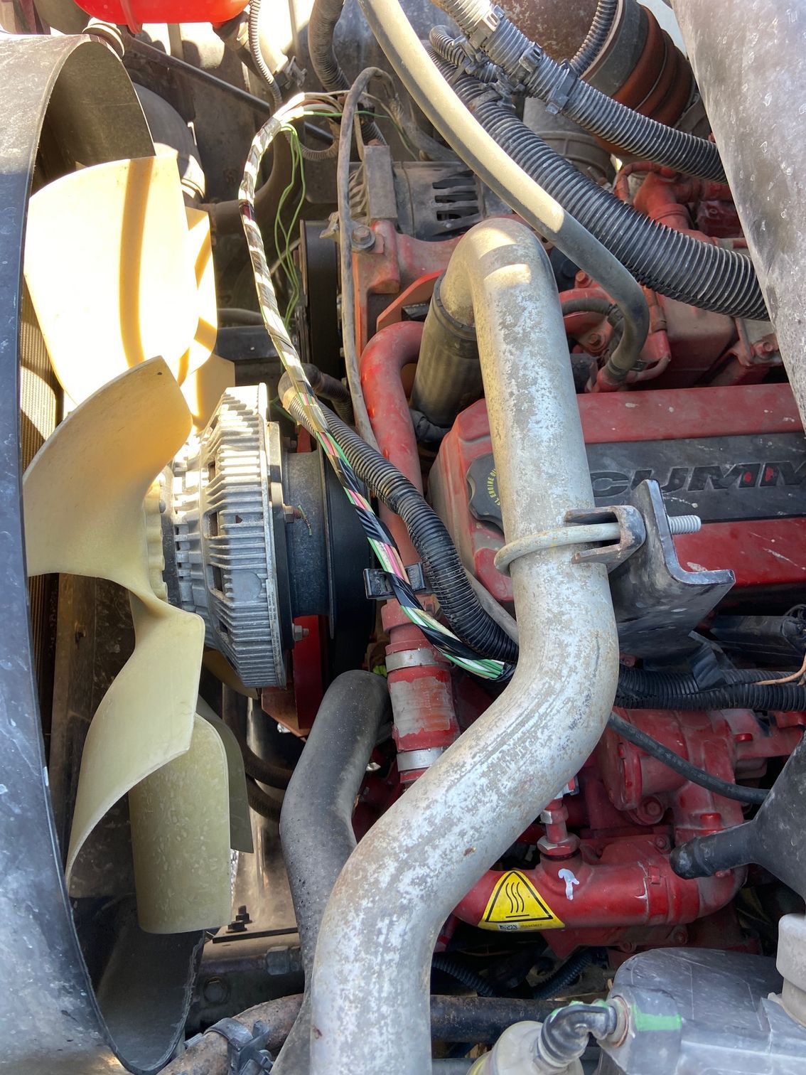 Engine bay of a truck, featuring a fan, belts, and various hoses and components. Red engine block visible.