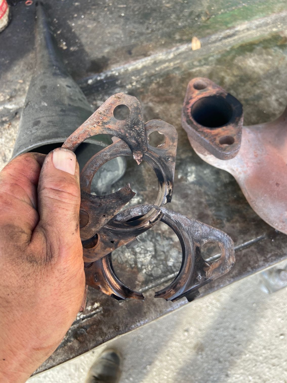 Hand holding damaged exhaust flange next to a rusted exhaust pipe.