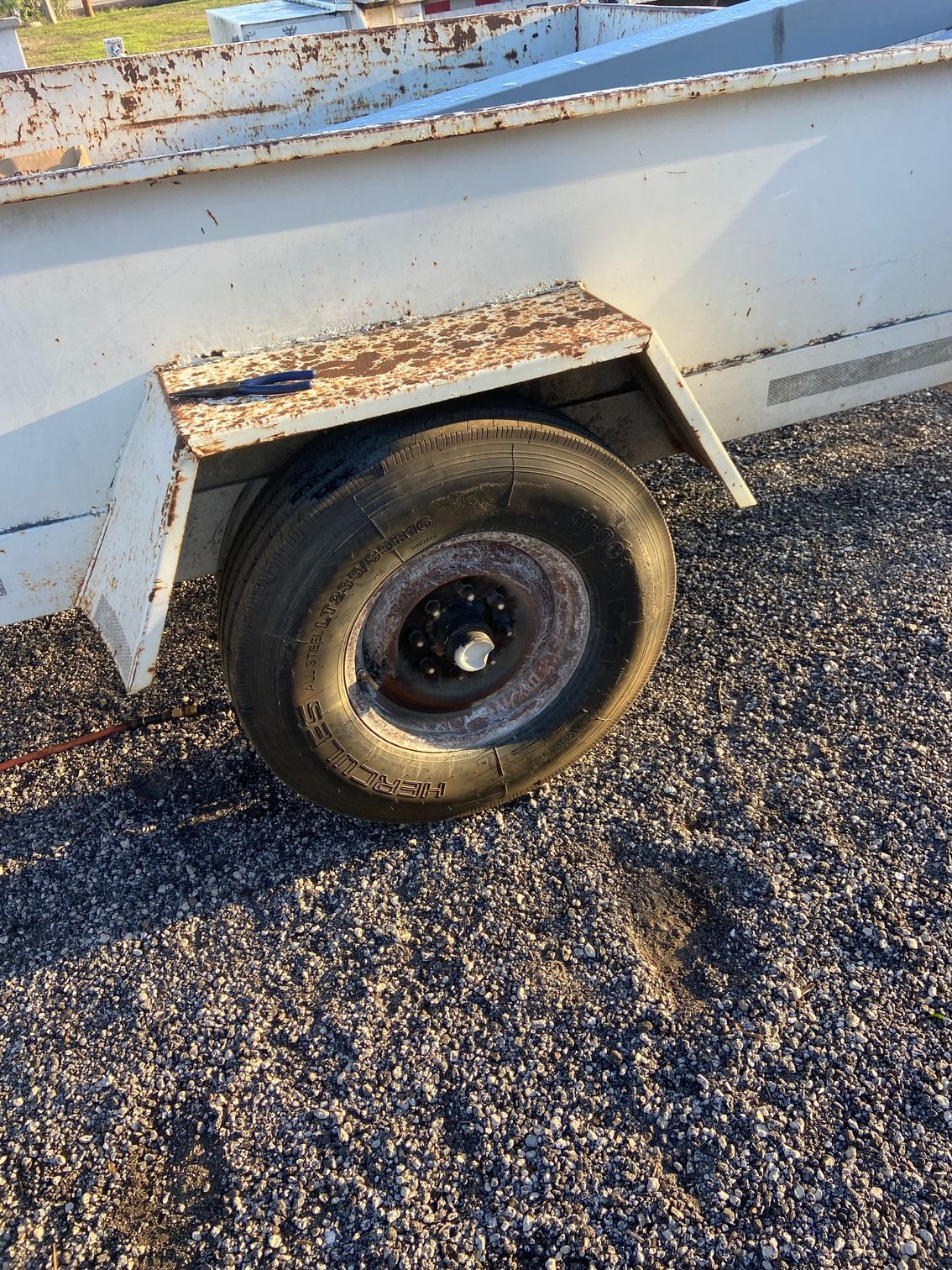 A light-colored trailer with a rusty fender and a black tire on a gravel surface.