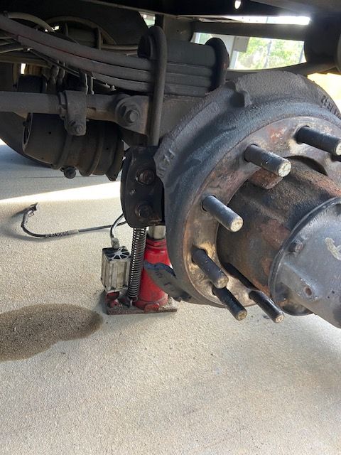 Truck's rear axle raised by a red hydraulic jack. Brake assembly and leaf spring visible. Gray pavement.
