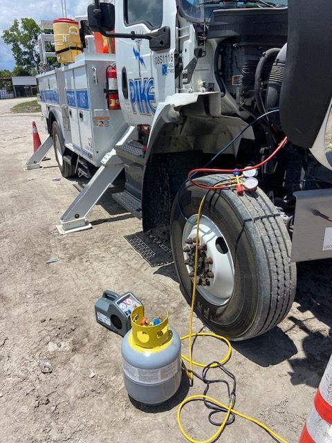 Truck being serviced; refrigerant tank, gauges, and hoses attached to tire. Gray and white truck. Outdoor setting.