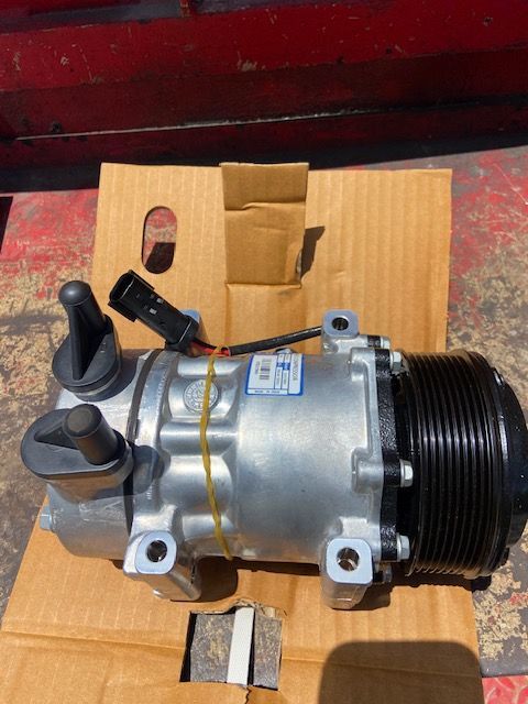 Silver and black air conditioning compressor in cardboard box on a red surface.