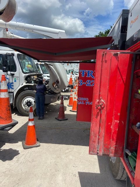 Person working on a white utility truck with an open hood, red tool cabinet, orange cones.