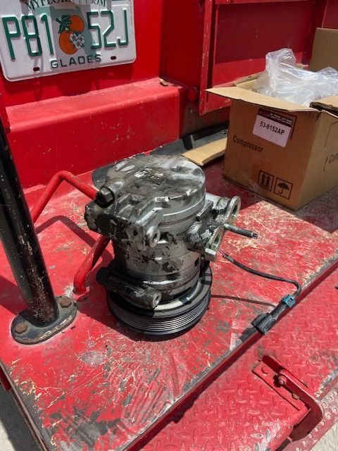 A car air conditioning compressor sits on a red surface next to a box and license plate.
